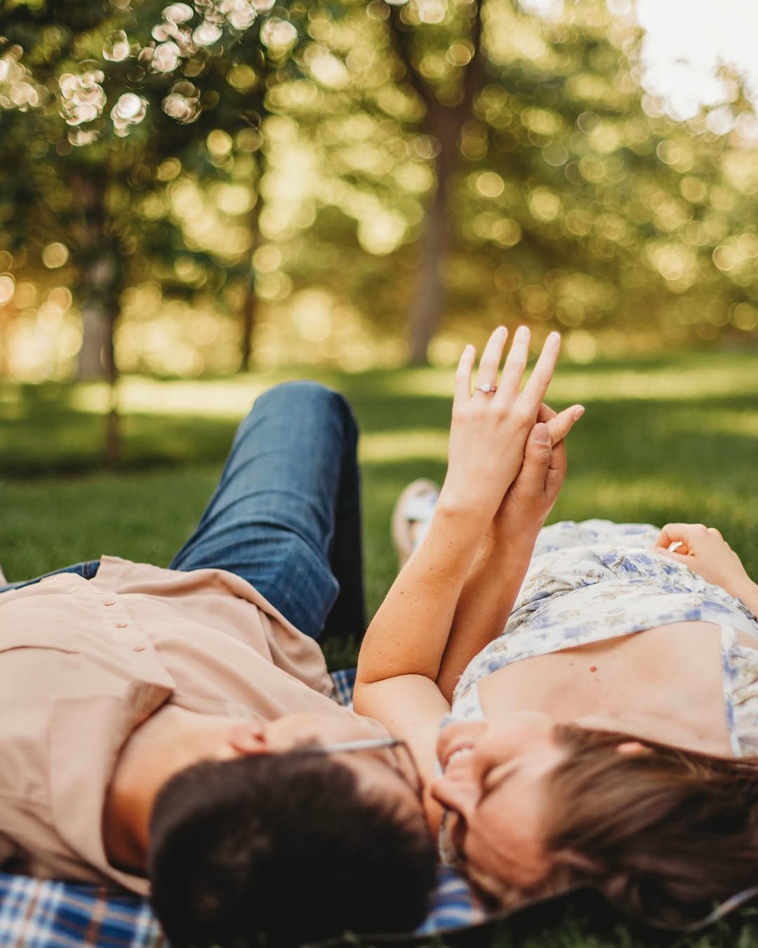 Couple lying on a picnic blanket in a sunny park, holding hands and laughing