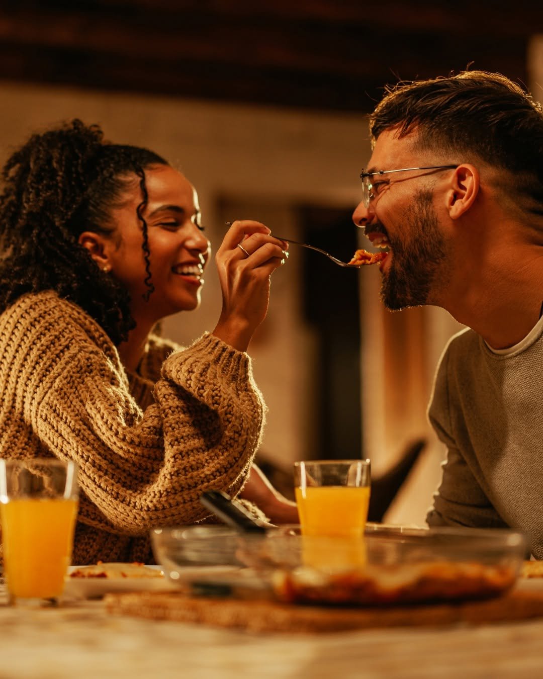 Couple laughing at dinner, woman playfully feeding her partner, from our date night trends guide