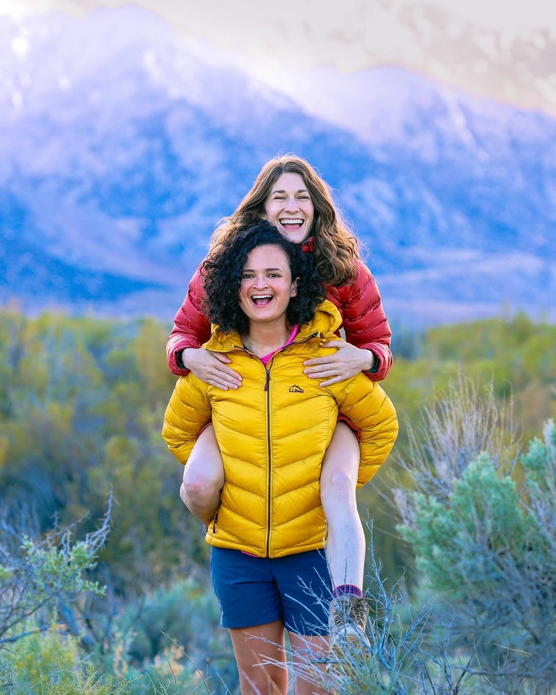 Couple outdoors with mountains behind, piggyback in hiking attire