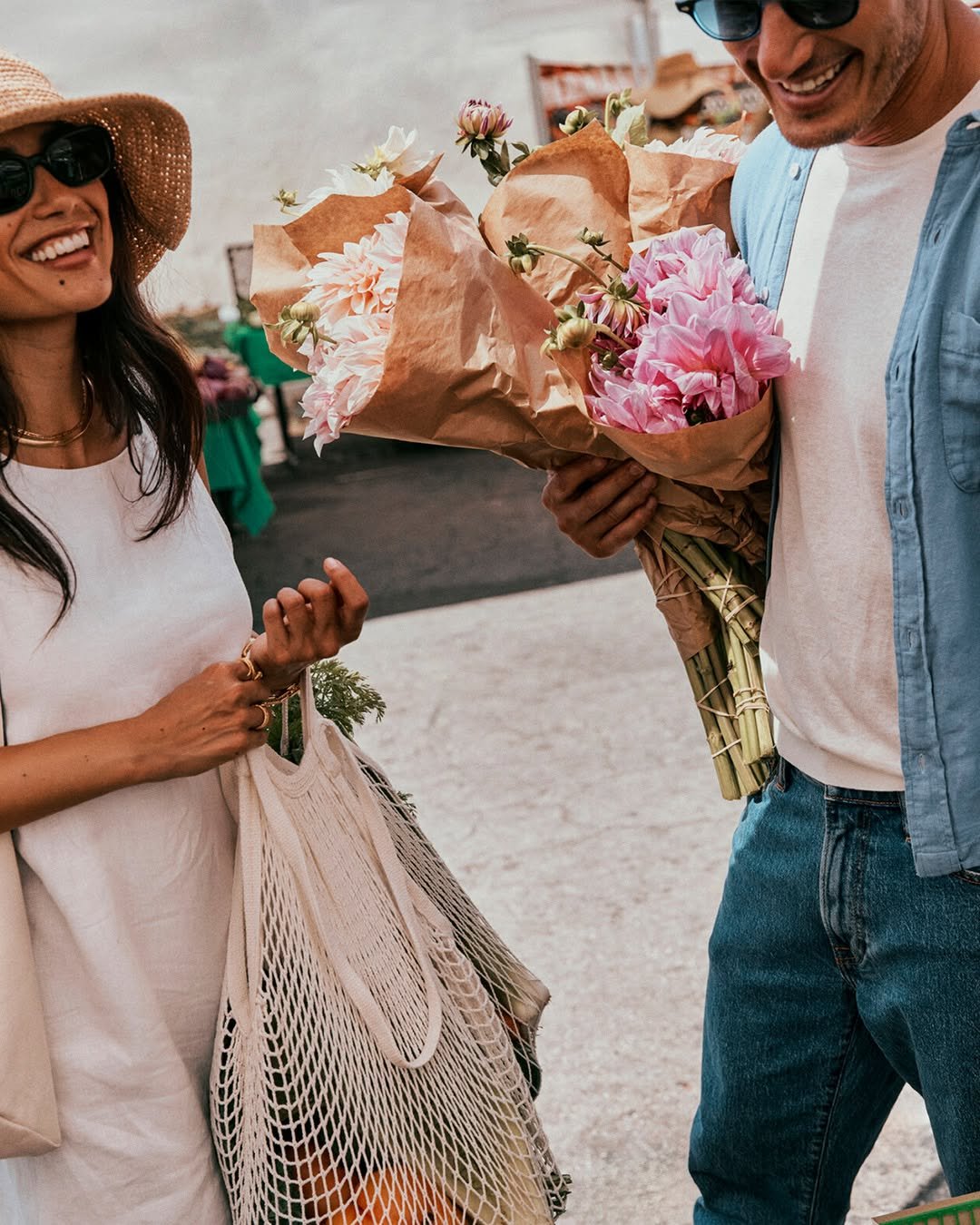 Smiling couple at a farmers market, woman with mesh bag, man with flowers