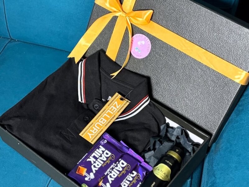 Black gift box with yellow ribbon holding polo shirt, chocolates and body spray on teal sofa
