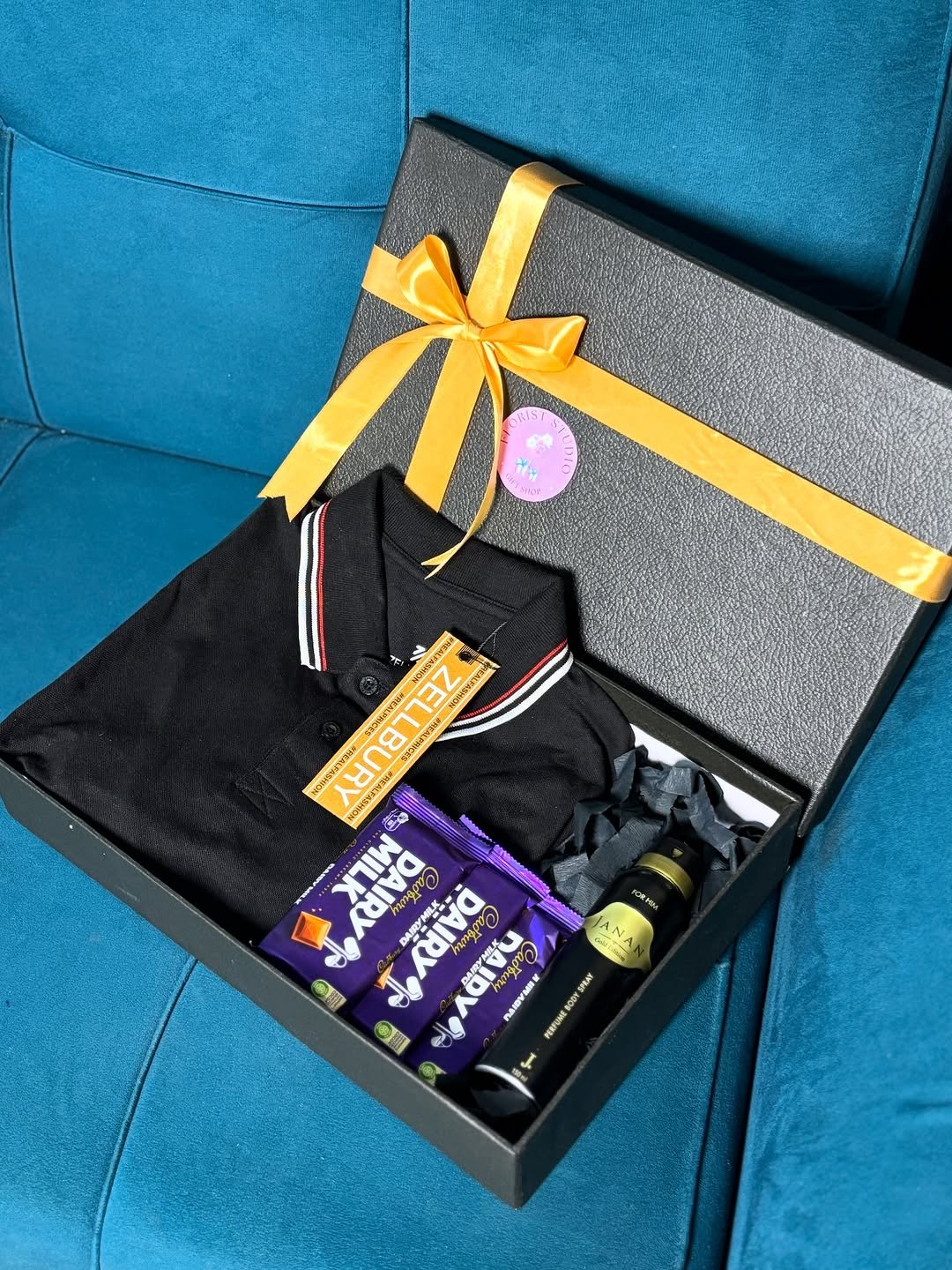 Black gift box with yellow ribbon holding polo shirt, chocolates and body spray on teal sofa