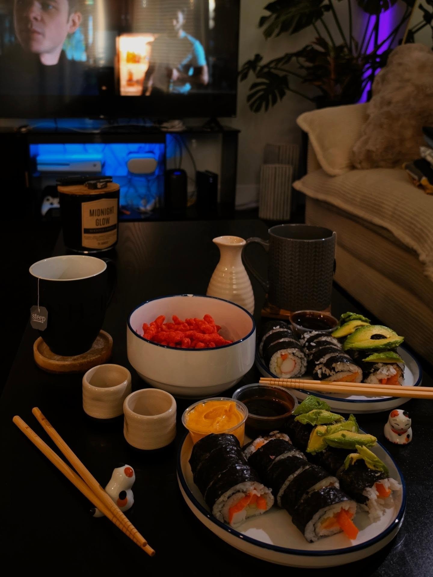 Sushi rolls and snacks spread on a coffee table before a glowing TV